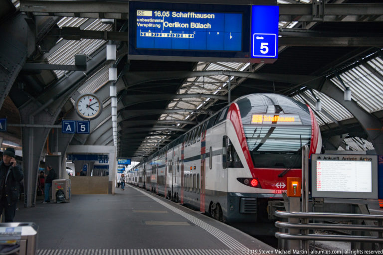 SBB double-deck train at a covered Zürich Hauptbahnhof platform beneath departure boards and an analog station clock.