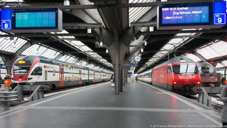 SBB double-deck train and red locomotive on adjacent Zürich Hauptbahnhof platforms beneath signs and a departure display.