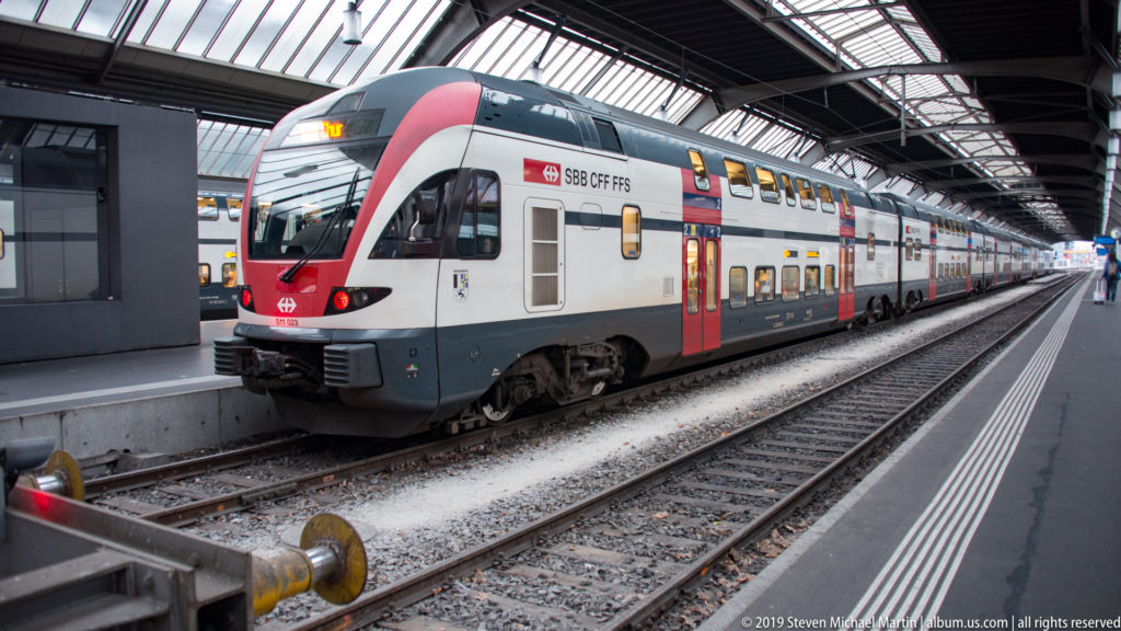 Red-and-white SBB double-deck passenger train standing beside a platform under Zürich Hauptbahnhof’s train shed.