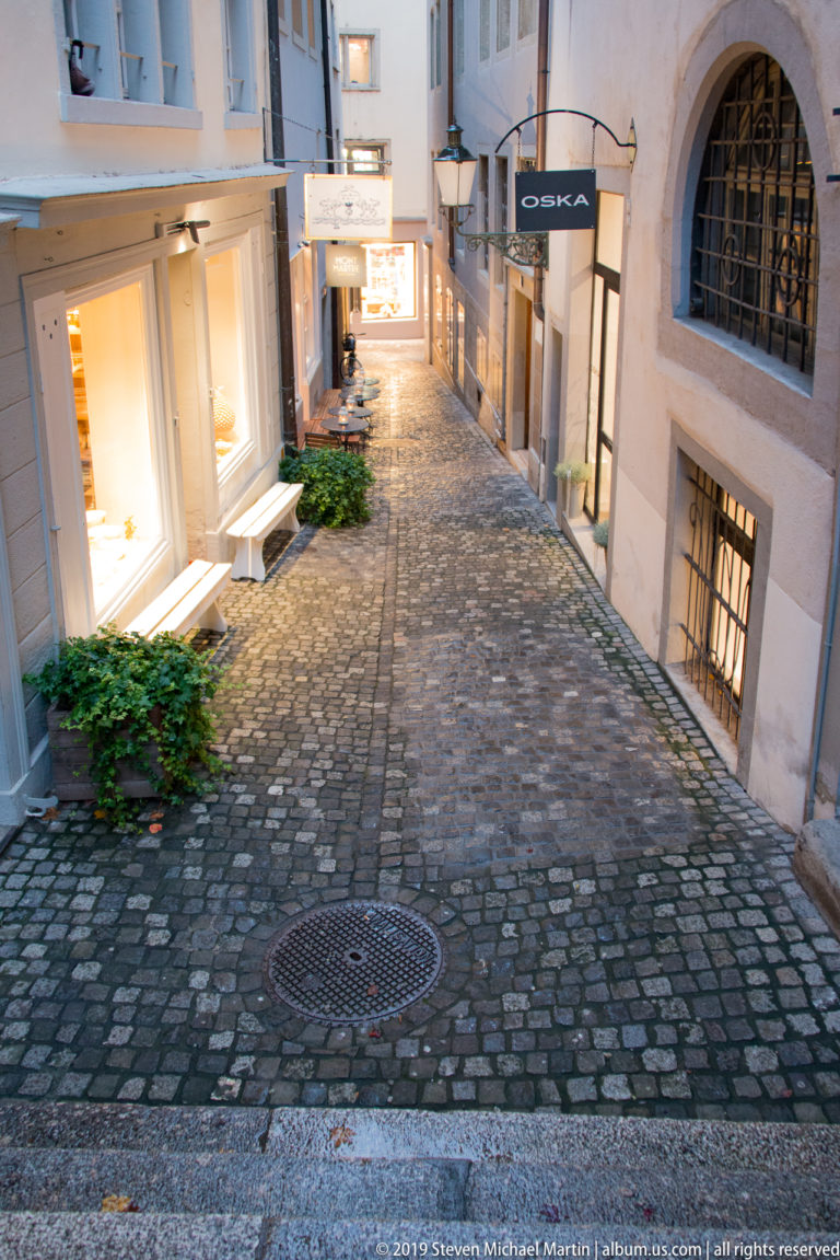 Empty cobblestone Old Town lane with glowing shop windows, barred windows, steps, and small patches of greenery.