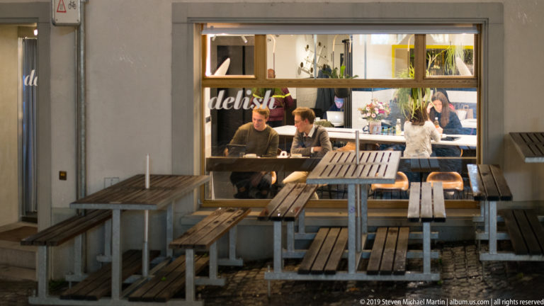 Empty wooden restaurant tables outside a brightly lit window where several people are visible inside.