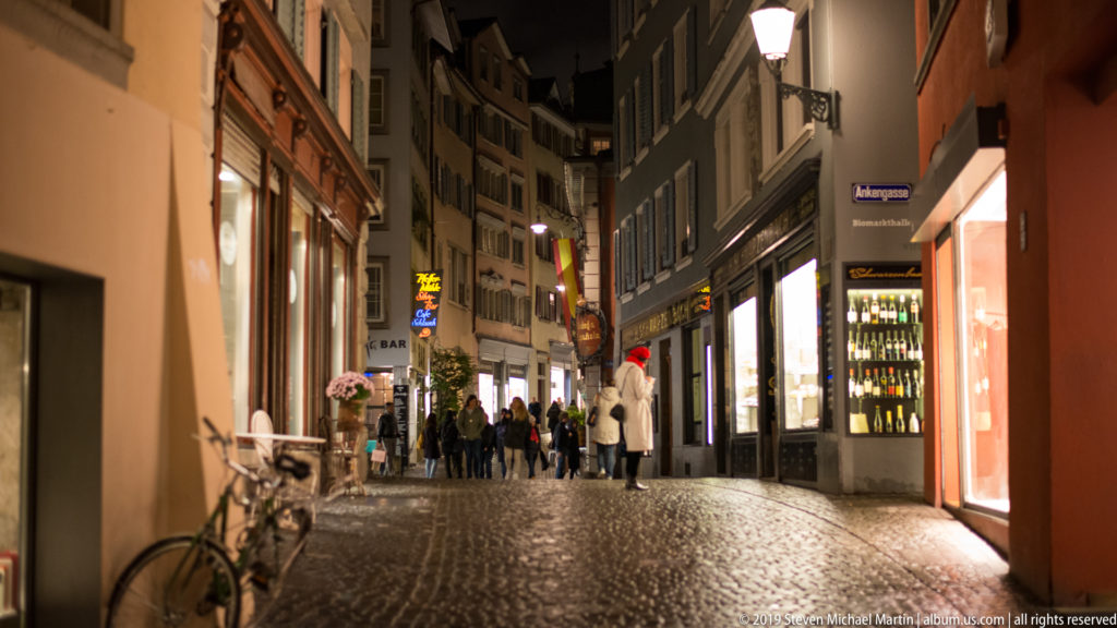 Narrow cobblestone Old Town street at night with bicycles, illuminated shops, pedestrians, and damp paving.