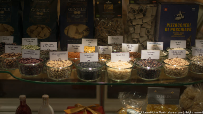 Glass containers of dried foods and seasonings beneath packaged pasta and specialty goods in a shop display.
