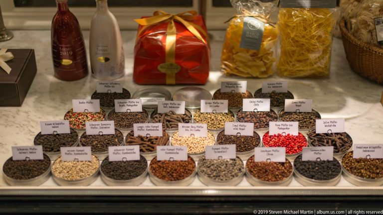 Rows of labeled white bowls filled with colorful spices, seeds, peppercorns, and seasoning blends.