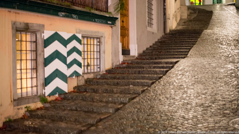 Narrow cobblestone lane and stone steps climbing past green-and-white chevron shutters in Zurich.