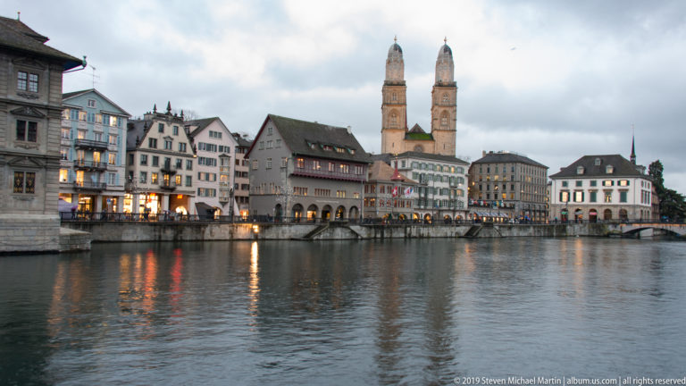 Wide evening view across the Limmat toward historic Zurich buildings and the twin towers of Grossmünster.