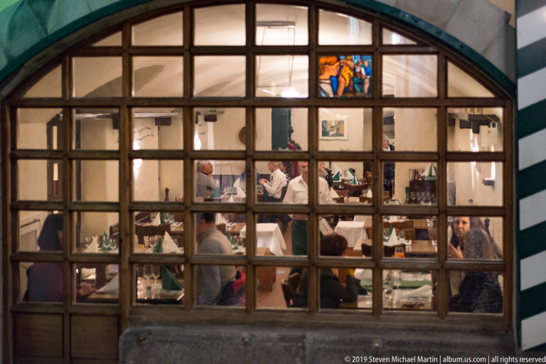 Diners seated at tables behind a large arched, multi-pane restaurant window at night.