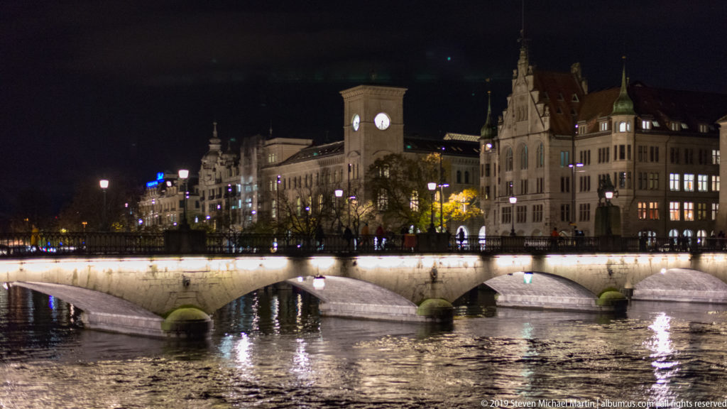 Illuminated stone arches of Münsterbrücke across the Limmat, with historic Limmatquai buildings and a clock tower beyond.