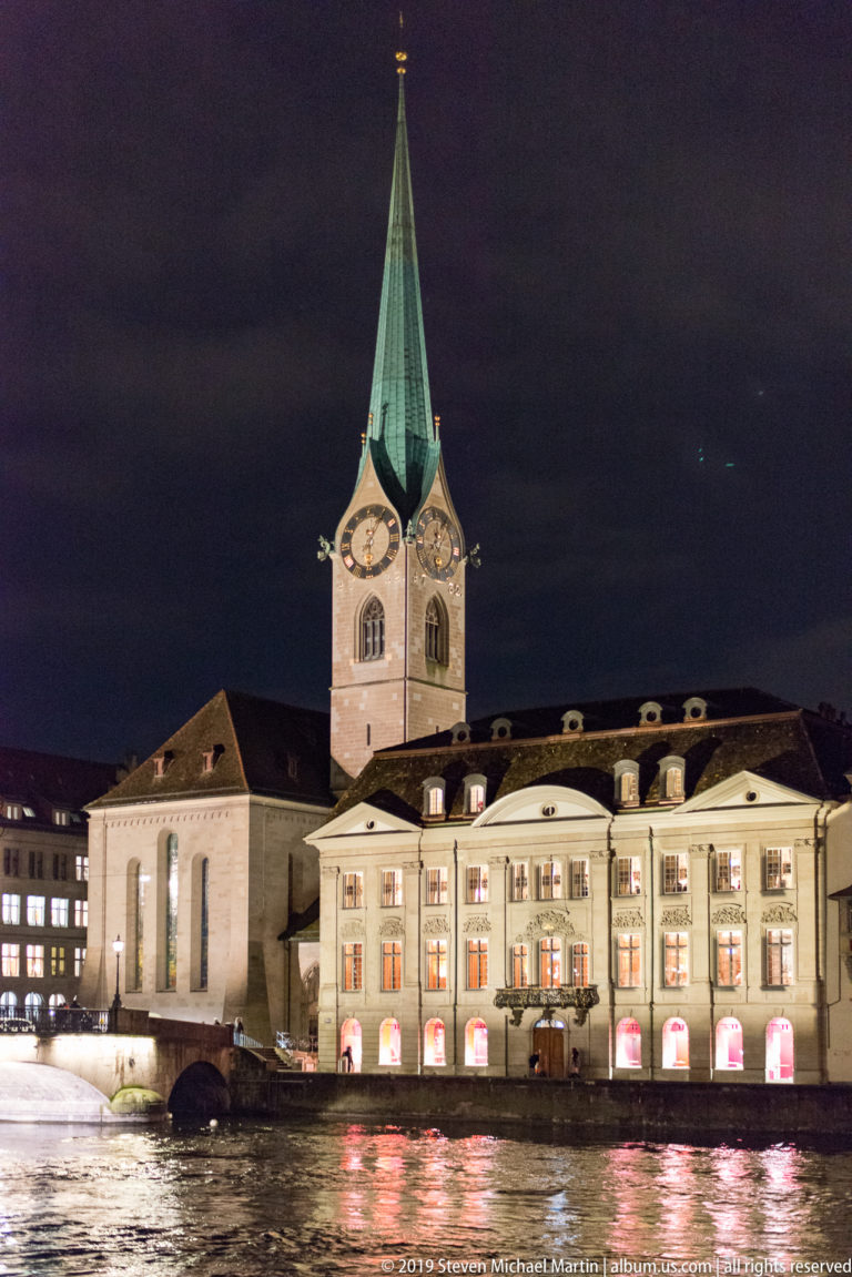 Fraumünster beside the Limmat at night, with its tall green spire above illuminated riverside buildings and reflections.