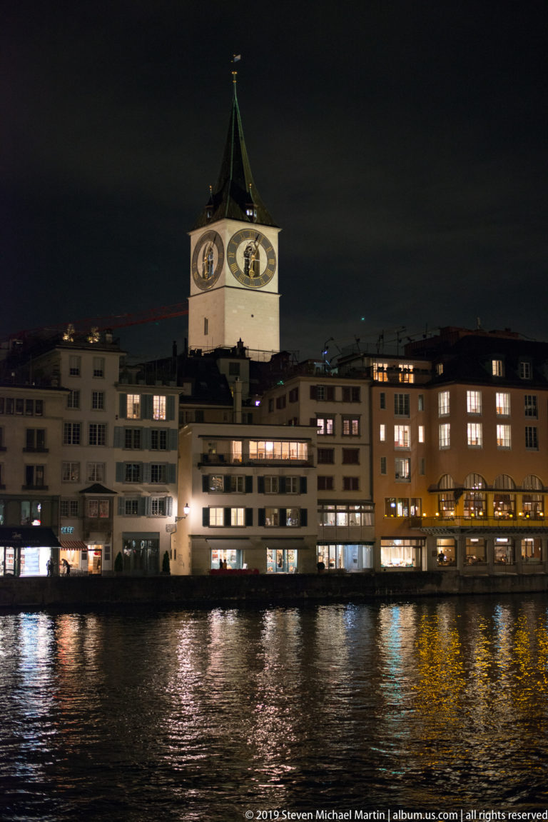St. Peter Church clock tower rising above waterfront buildings across the Limmat, with colored reflections on the river.