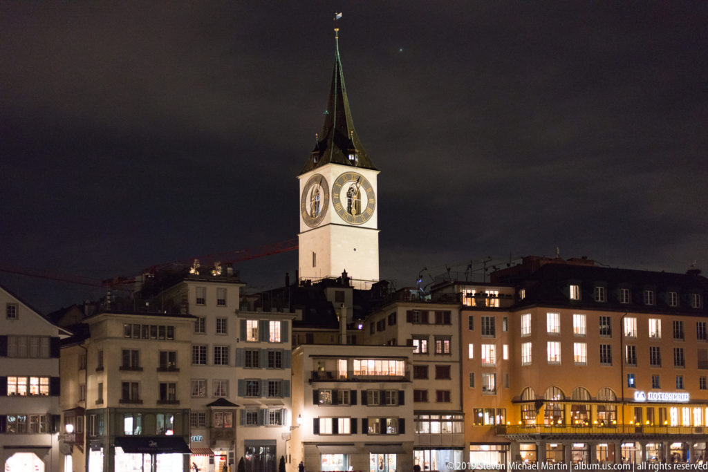 St. Peter Church clock tower above illuminated Zurich Old Town buildings, with large clock faces and a dark green spire.
