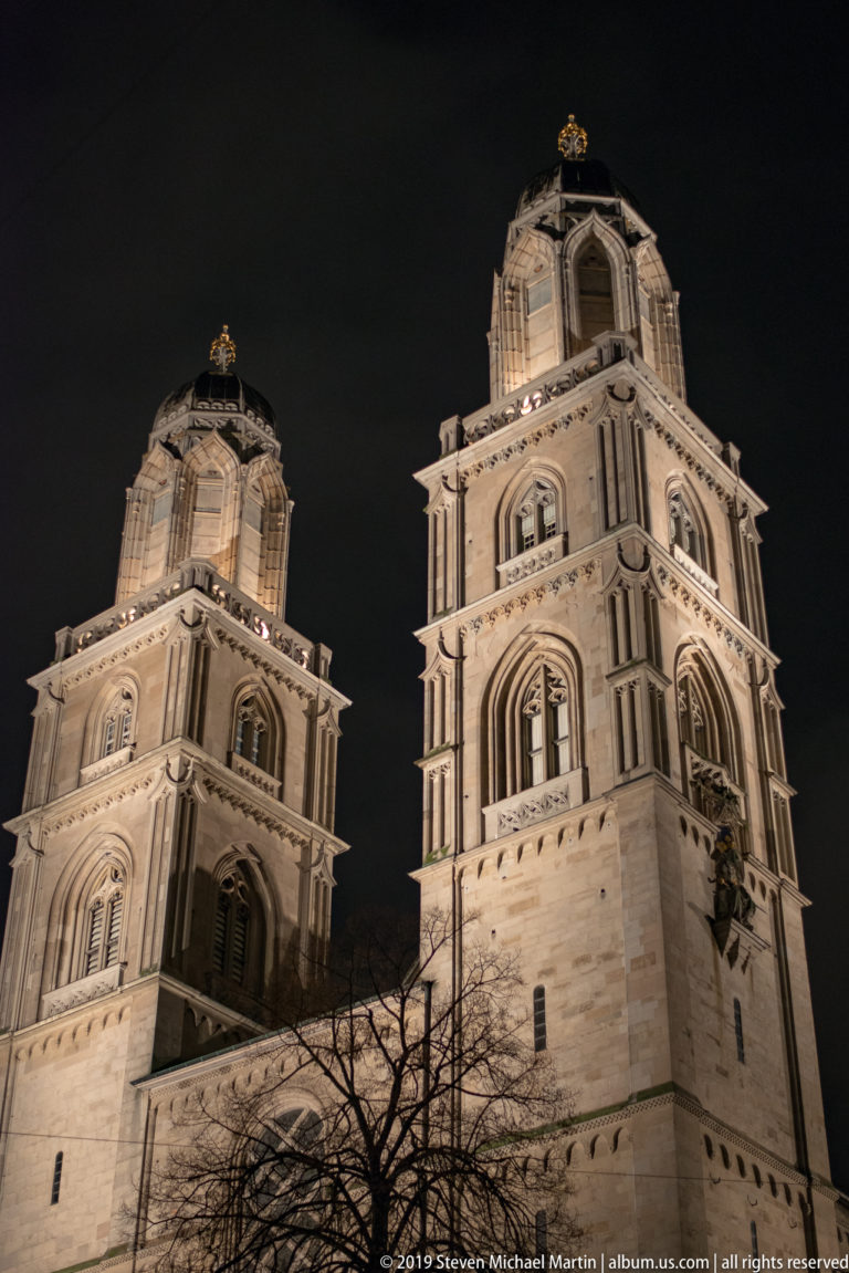 Low-angle night view of the illuminated twin Grossmünster towers rising above bare tree branches.