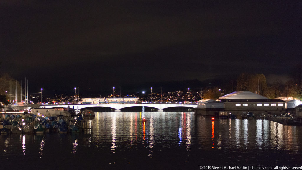 Dark Limmat River reflecting bridge and waterfront lights, with low buildings and boat masts along the banks.