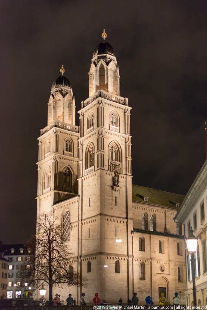 Close view of the illuminated twin towers of Grossmünster at night, with a bare tree and lower rooftops below.