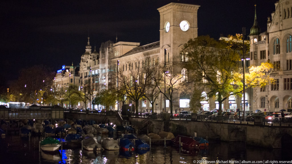Moored boats on the Limmat below illuminated historic buildings and a square clock tower along Zurich’s waterfront.