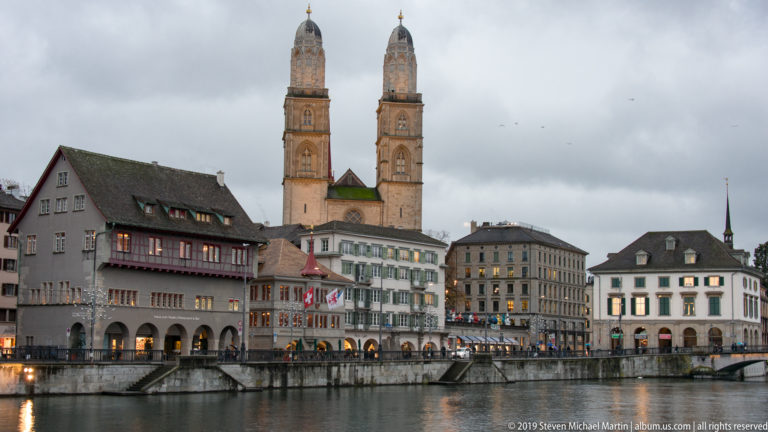 Grossmünster twin towers above historic buildings along the Limmat, with evening reflections on the river.