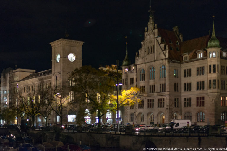 Historic buildings and a square clock tower along the Limmatquai at night, with moored boats in the foreground.