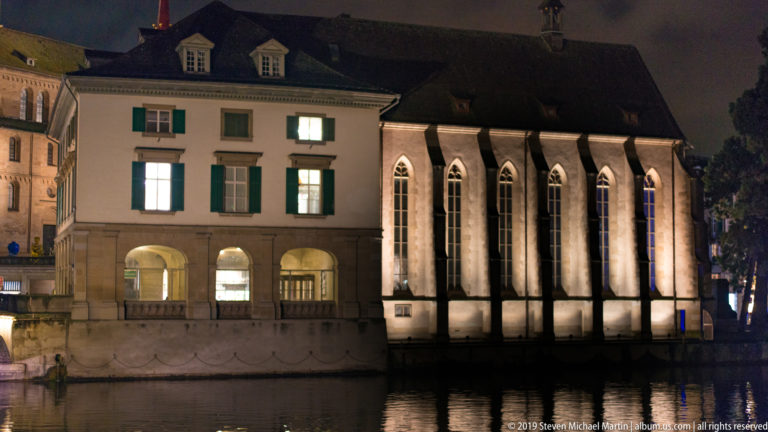 Wasserkirche along the Limmat at night, with tall pointed windows beside a pale building with green shutters.