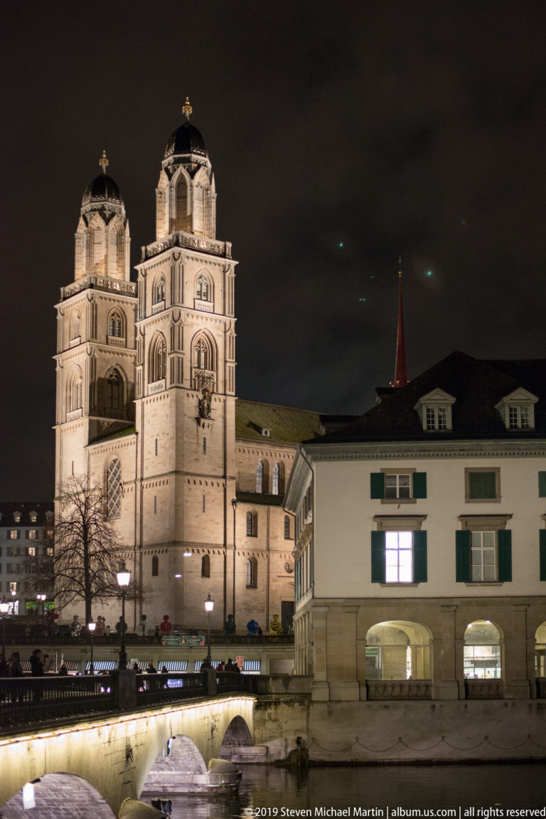 Illuminated twin towers of Zurich’s Grossmünster above riverside buildings and a lit bridge at night.
