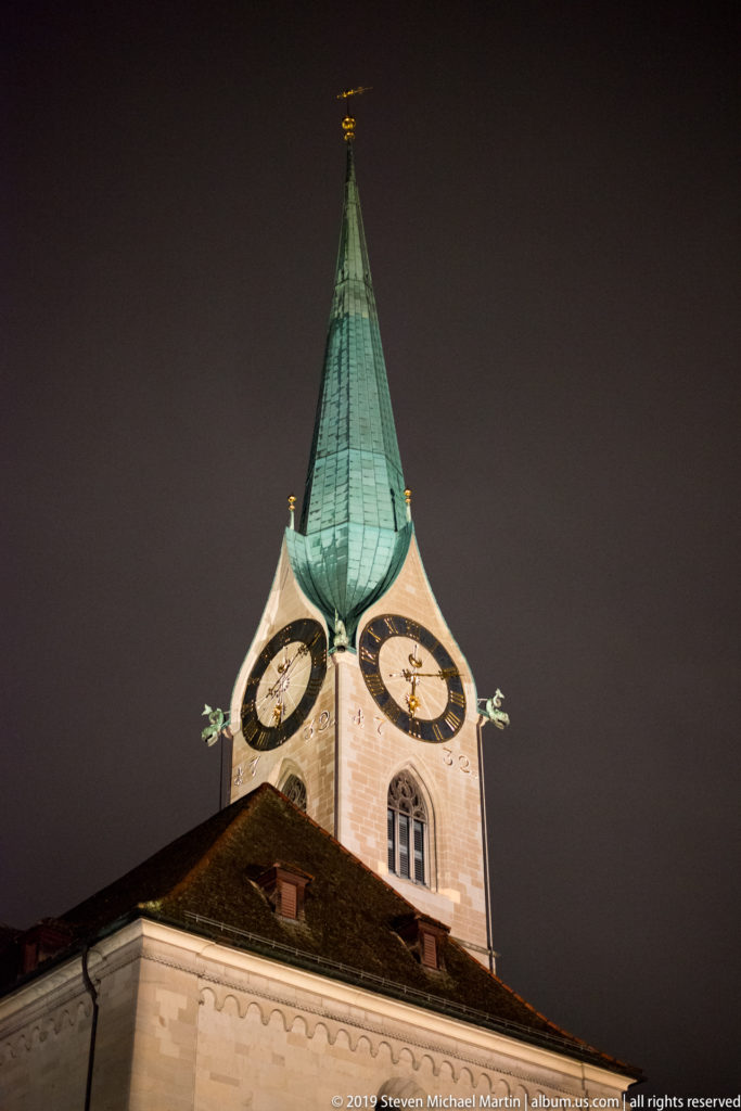 St. Peter Church tower in Zurich at night, with two large clock faces and a tall green copper spire.