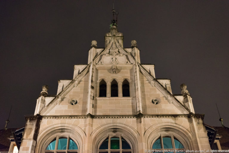 Symmetrical illuminated stone gable with three tall arched windows, circular openings, finials, and a small upper arcade.