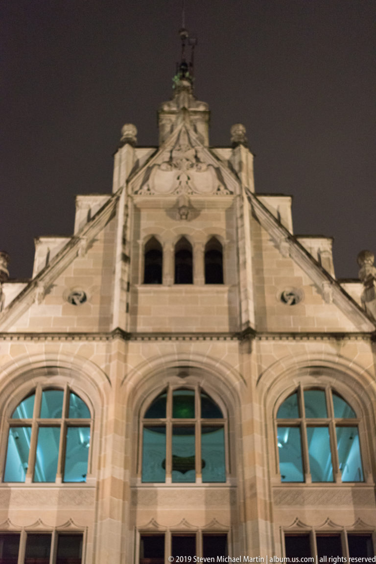 Illuminated pale-stone gabled façade with three large arched windows, circular openings, and a triple-arched opening near the peak.
