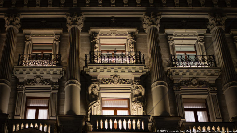Ornate historic façade at night with tall columns, carved window surrounds, balconies, balustrades, and sculptural figures.