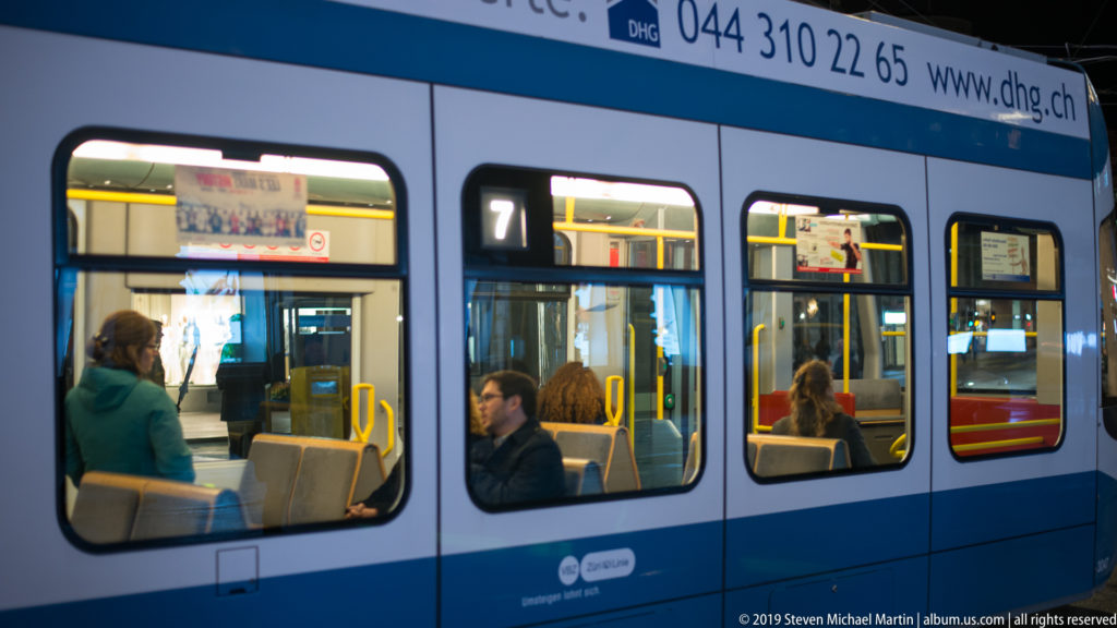 Blue-and-white Zurich route 7 tram at night, with seated passengers visible through illuminated windows.