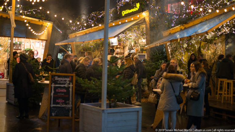 Crowd in winter coats gathered around a brightly lit Glühwein stall with evergreen decorations at night.