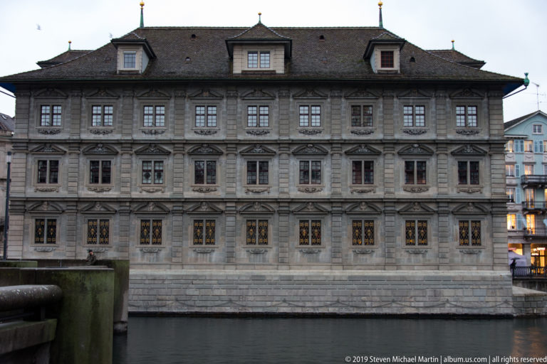 Zurich Rathaus seen frontally across the Limmat, with symmetrical stone façade, rows of windows, and steep roof.