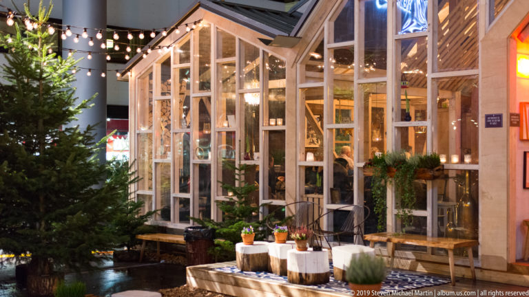 Illuminated glass-and-wood pavilion beside a fir tree, with plants, tables, and small displays visible inside and outside.