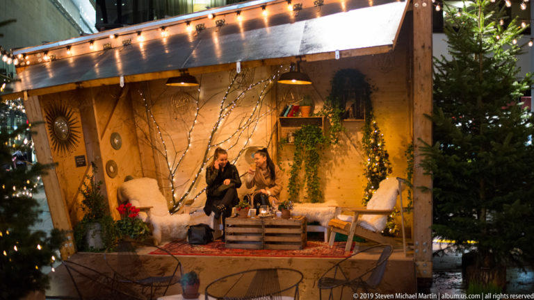 Two people seated in a warmly lit timber Christmas-market hut decorated with greenery, branches, lamps, and cushions.