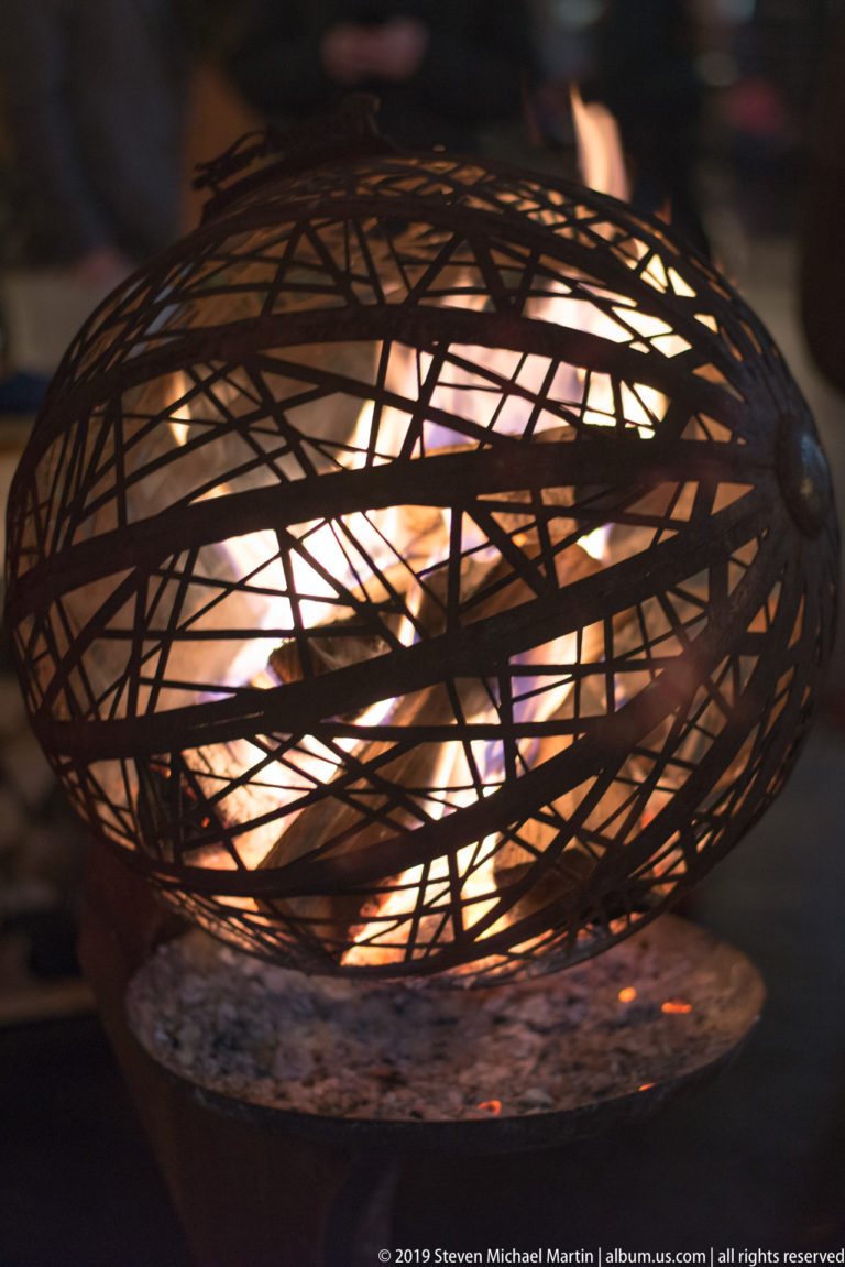 Flames glowing inside a spherical basket of interwoven dark metal strips at night.