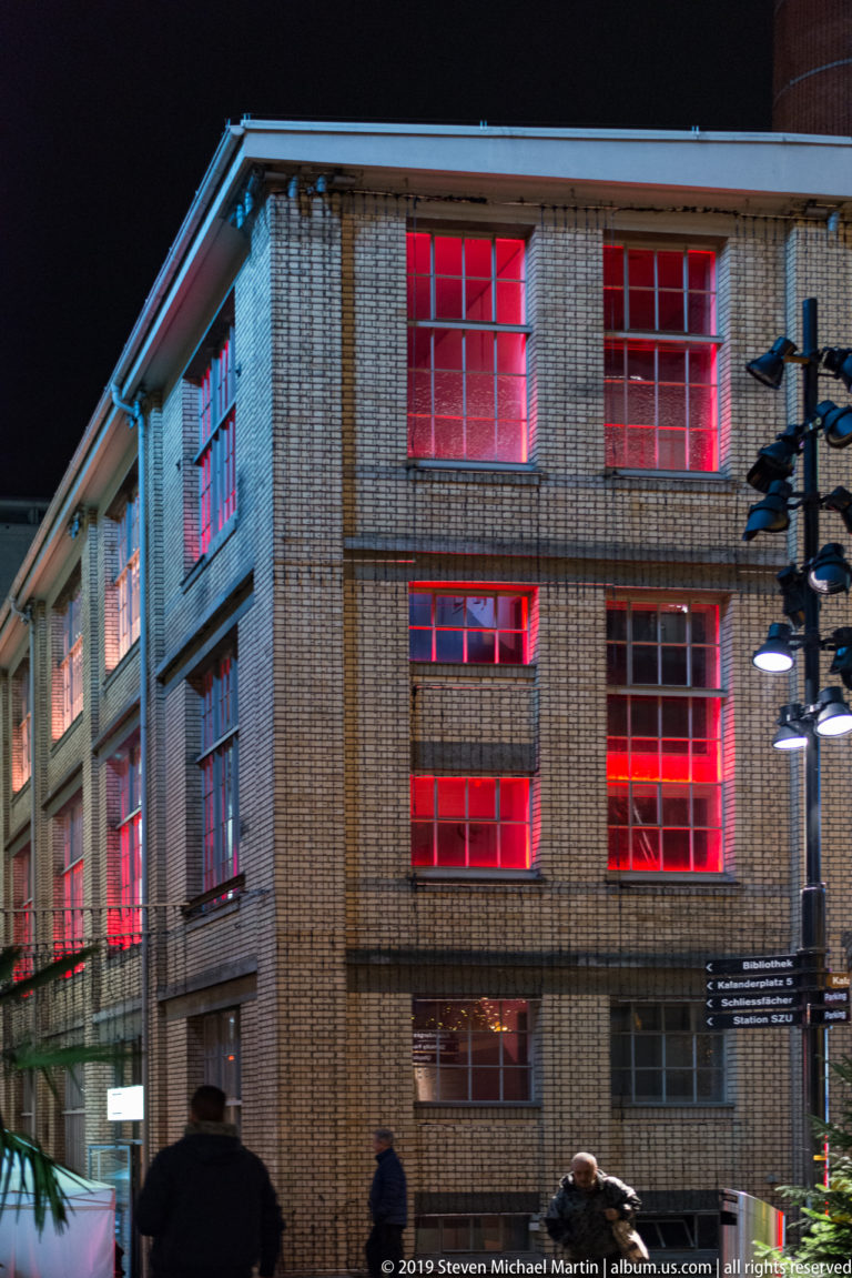 Large brick industrial-style building with red-lit windows and pedestrians along the street at night.