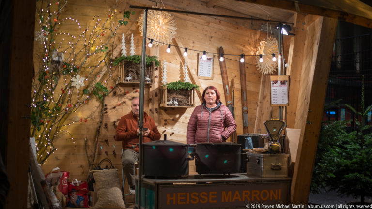 Vendor inside a wooden stall marked “Heisse Marroni,” with holiday decorations, branches, and chestnut-roasting equipment.