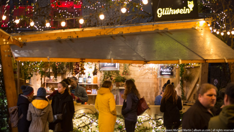 People in winter coats gather at a timber Glühwein stall decorated with lights and greenery at a Zurich Christmas market.