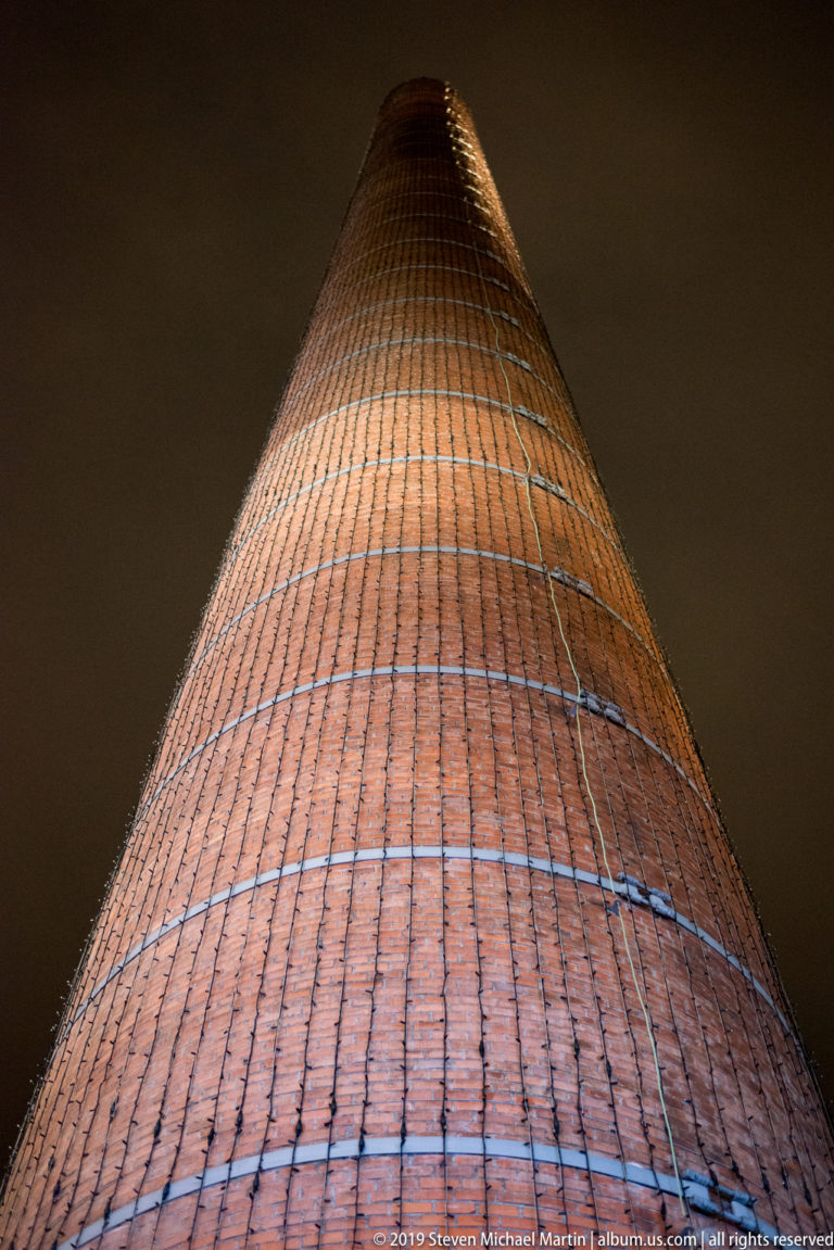 Upward view of a tall tapered brick chimney wrapped with horizontal metal bands against the night sky.