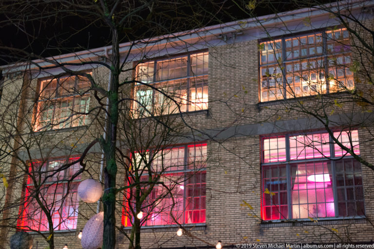 Brick industrial-style building with large windows glowing red and pink behind bare tree branches at night.