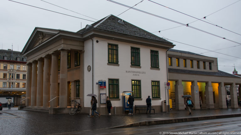 Kantonspolizei Zürich building with columned portico, wet pavement, overhead tram wires, pedestrians, and bicycles.