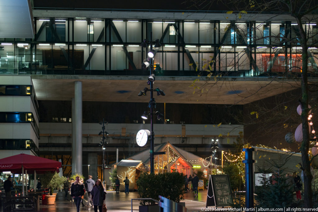 Christmas market stalls and visitors beneath a modern concrete-and-glass building in Zurich at night.