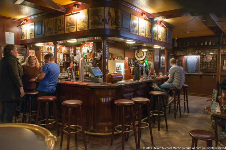 Curved wooden pub bar with stools, beer taps, bottles, framed decorations, and patrons gathered around it.