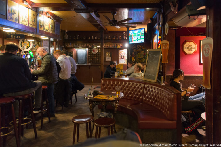 Warm wood-paneled pub interior with patrons at the bar, leather banquettes, stools, beer signs, and ceiling fans.