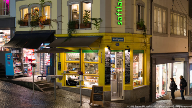 Bright corner shop at Marktgasse 31 with yellow-green façade, awnings, illuminated displays, and pedestrians nearby.