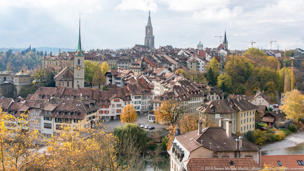 Wide Bern cityscape with red roofs, church spires and Bern Minster tower above the Aare valley.