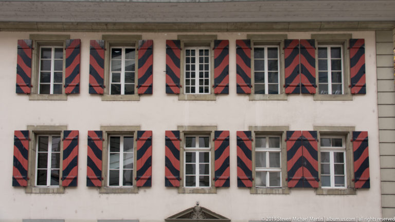 Historic pale façade with rows of windows and red-and-black geometric shutters.