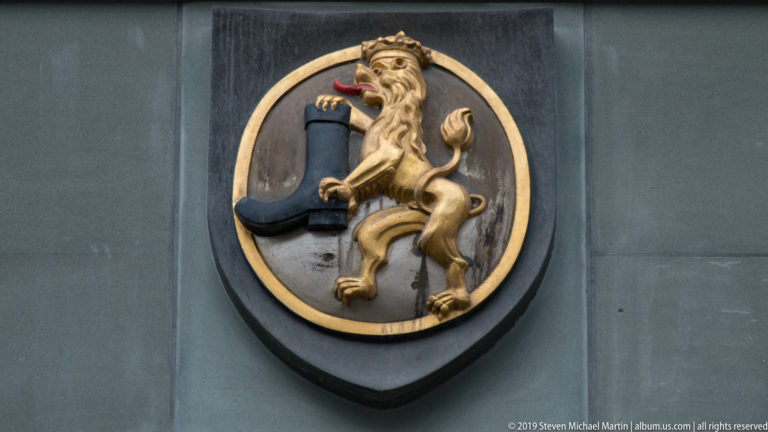 Gilded crowned lion beside a blue horn emblem on a black oval heraldic plaque.
