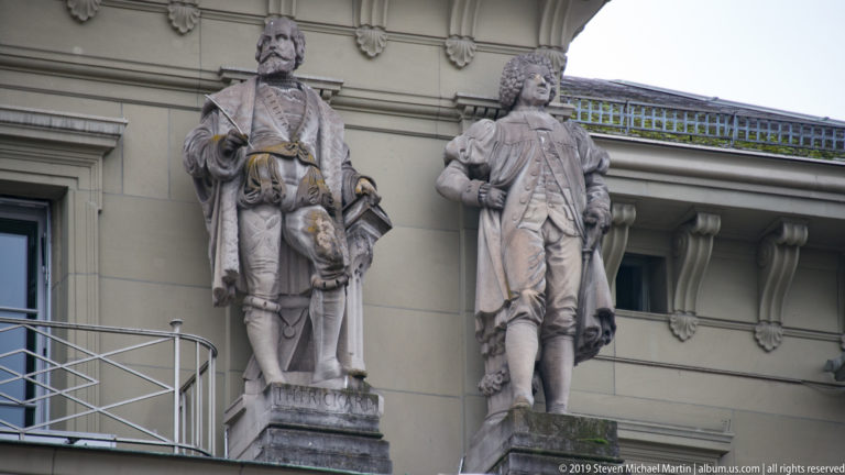 Two stone statues of men in historical dress mounted on the Swiss Federal Palace façade.