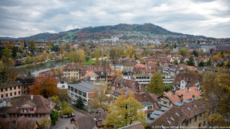 Elevated view over Bern rooftops and autumn trees toward a densely settled wooded hill.