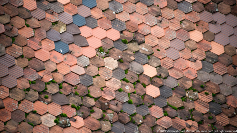 Close view of weathered overlapping clay roof tiles in varied red, brown, gray, and black tones.