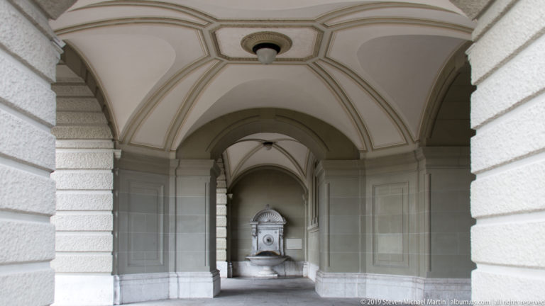 Vaulted stone passage with repeating arches leading to a wall fountain in the Swiss Federal Palace complex.